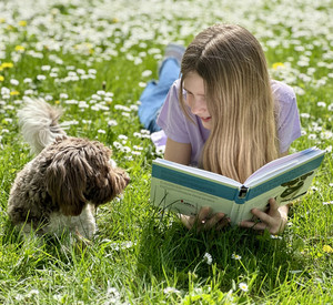 Lachendes blondes Mädchen liegt neben Hund auf der Wiese und hält ein Buch in den Händen