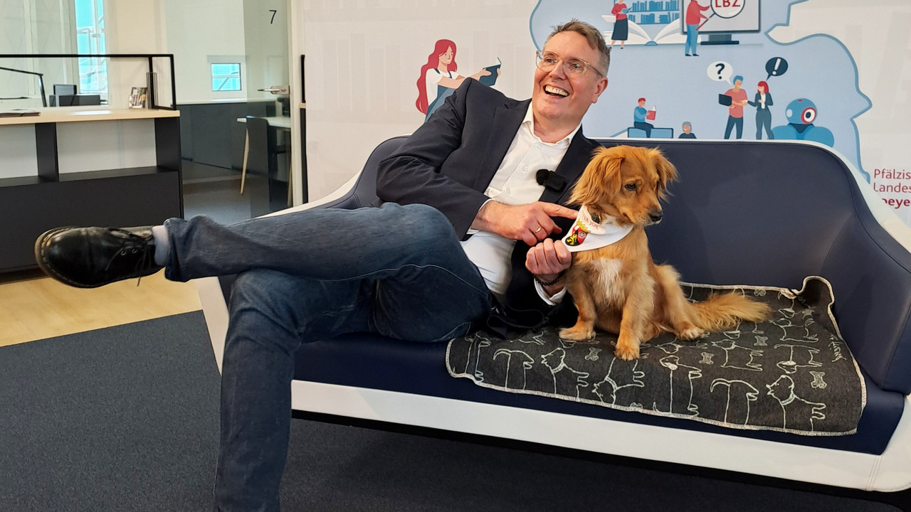 Ministerpräsident Alexander Schweitzer sitzt lachend auf einer Couch mit Hund Little Joe.