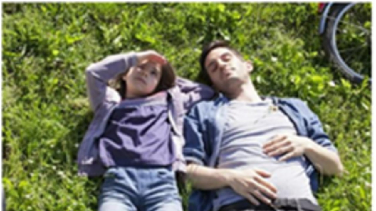 Zwei Personen, die auf einer grünen Wiese auf dem Rücken liegen und in den Himmel schauen, Sie sind in grau-blau Töne gekleidet. Oben rechts ragt das Rad eines Fahrrads ins Bild