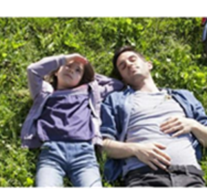 Zwei Personen, die auf einer grünen Wiese auf dem Rücken liegen und in den Himmel schauen, Sie sind in grau-blau Töne gekleidet. Oben rechts ragt das Rad eines Fahrrads ins Bild