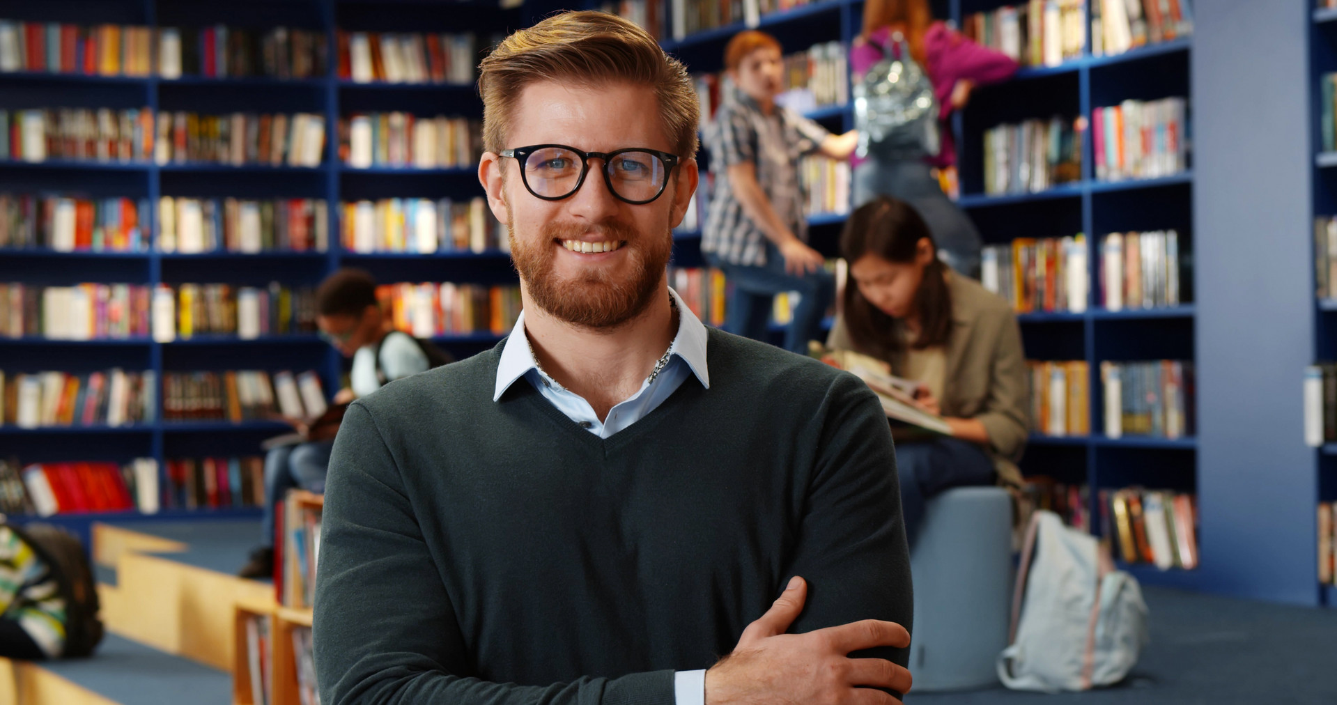 Portrait of young male teacher n library with other students studying in background. Handsome librarian smiling at camera standing in public school library