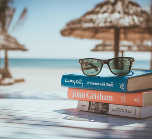 Sonnenbrille auf Bücherstapel, Im Hintergrund: Ein weißer Strand mit Sonnenschirmen